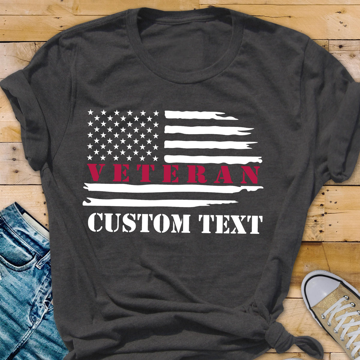 May include: Dark gray t-shirt featuring a distressed American flag design. The flag includes white stars and stripes, with the word "VETERAN" in red and "CUSTOM TEXT" in white. The shirt is a casual style.