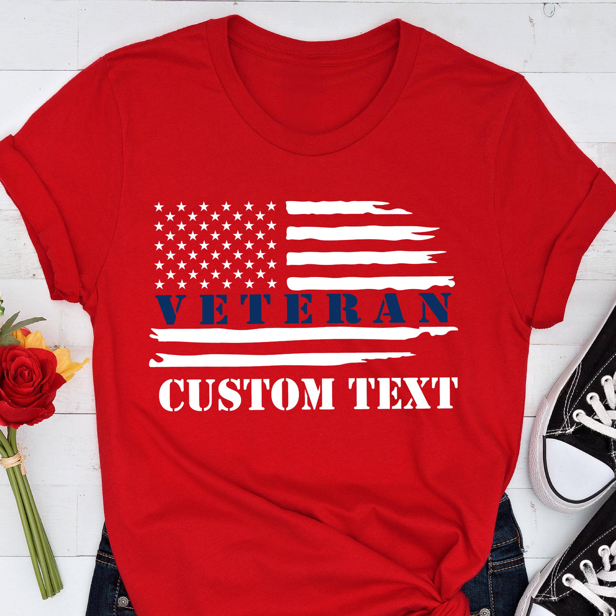 May include: Red t-shirt featuring a distressed American flag design. The flag includes white stars and stripes, with the word "VETERAN" in blue and space for custom text in white. The shirt is paired with dark blue jeans and black sneakers.