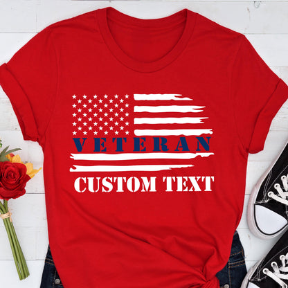 May include: Red t-shirt featuring a distressed American flag design. The flag includes white stars and stripes, with the word "VETERAN" in blue and space for custom text in white. The shirt is paired with dark blue jeans and black sneakers.
