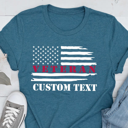 May include: Teal t-shirt featuring a distressed American flag design. The word "VETERAN" is printed in red across the flag, with the words "CUSTOM TEXT" below in white. A casual, patriotic apparel item.