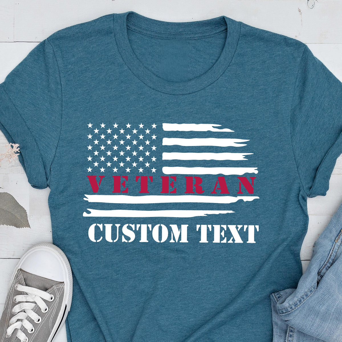 May include: Teal t-shirt featuring a distressed American flag design. The word "VETERAN" is printed in red across the flag, with the words "CUSTOM TEXT" below in white. A casual, patriotic apparel item.