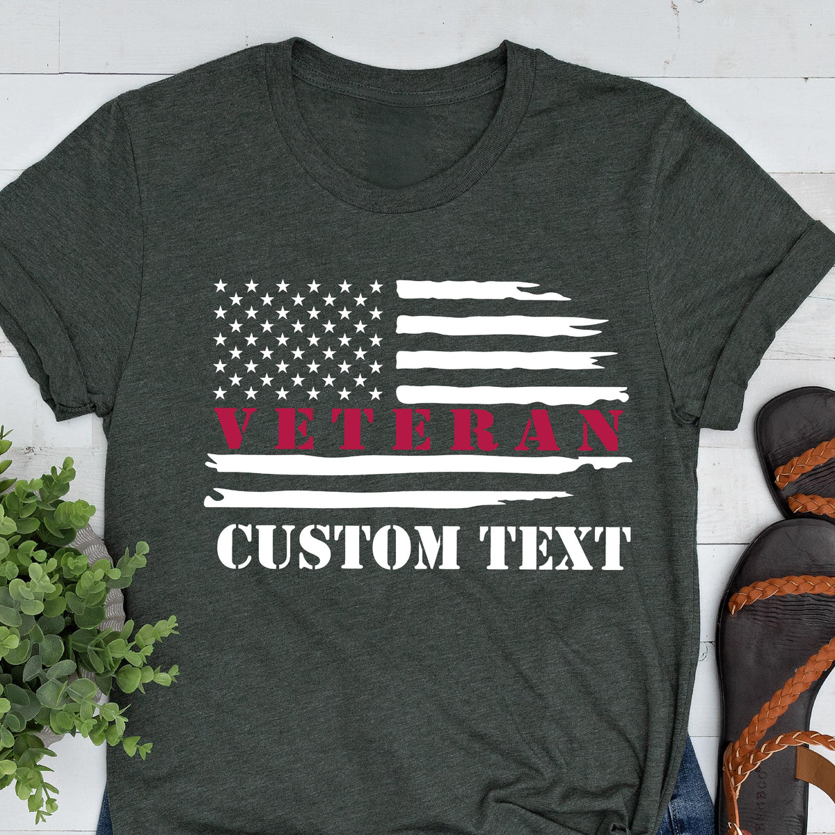 May include: Dark gray t-shirt featuring a distressed American flag design. The flag incorporates white stars and stripes, with the word "VETERAN" in red. Below the flag is the text "CUSTOM TEXT" in white, allowing for personalization.