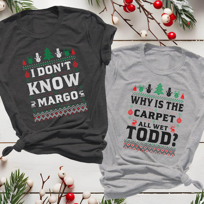 May include: Two Christmas-themed t-shirts. One is dark gray with the text "I DON'T KNOW MARGO" in white, red, and green. The other is light gray with "WHY IS THE CARPET ALL WET TODD?" in similar colors. Both shirts have festive patterns.