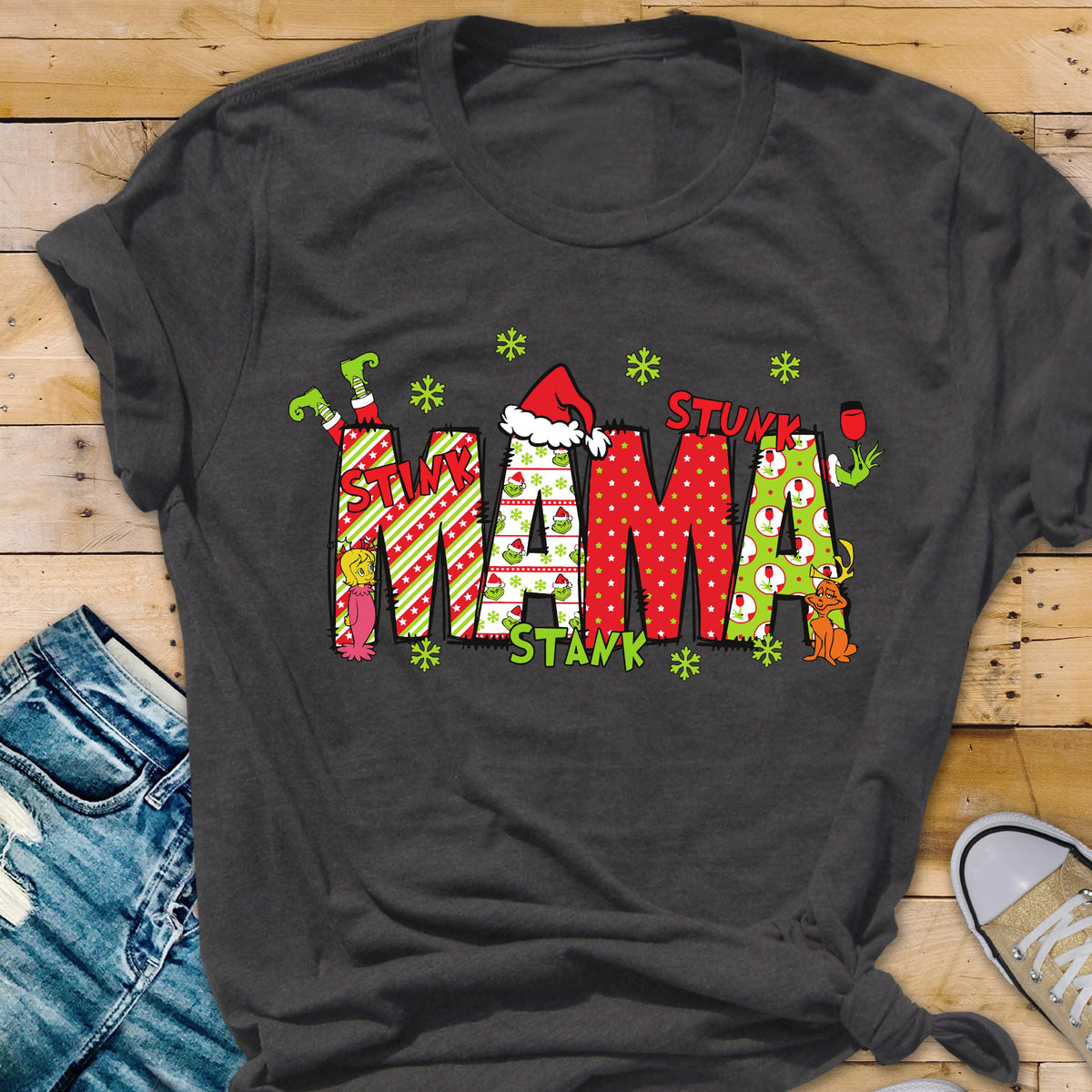May include: A dark gray short-sleeve t-shirt featuring the word MAMA in large, patterned block letters. The letters include red and white stripes, green and white snowflakes, and red polka dots. The design includes a Santa hat and green characters.