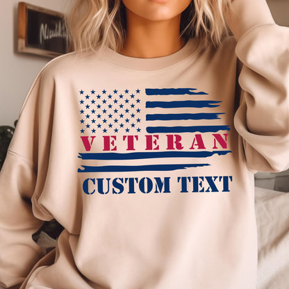 May include: Beige sweatshirt featuring a distressed American flag design. The flag includes a field of blue stars, red and blue stripes, and the word "VETERAN" in red. Below the flag is the text "CUSTOM TEXT" in blue.