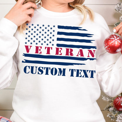 May include: White sweatshirt featuring a distressed American flag design with the word "VETERAN" in red and "CUSTOM TEXT" in navy blue. The sweatshirt has a crew neck and long sleeves.