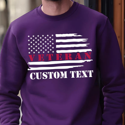 May include: A purple sweatshirt featuring a distressed American flag design. The flag includes white stars and stripes, with the word "VETERAN" in red and "CUSTOM TEXT" in white. The sweatshirt is a solid color.