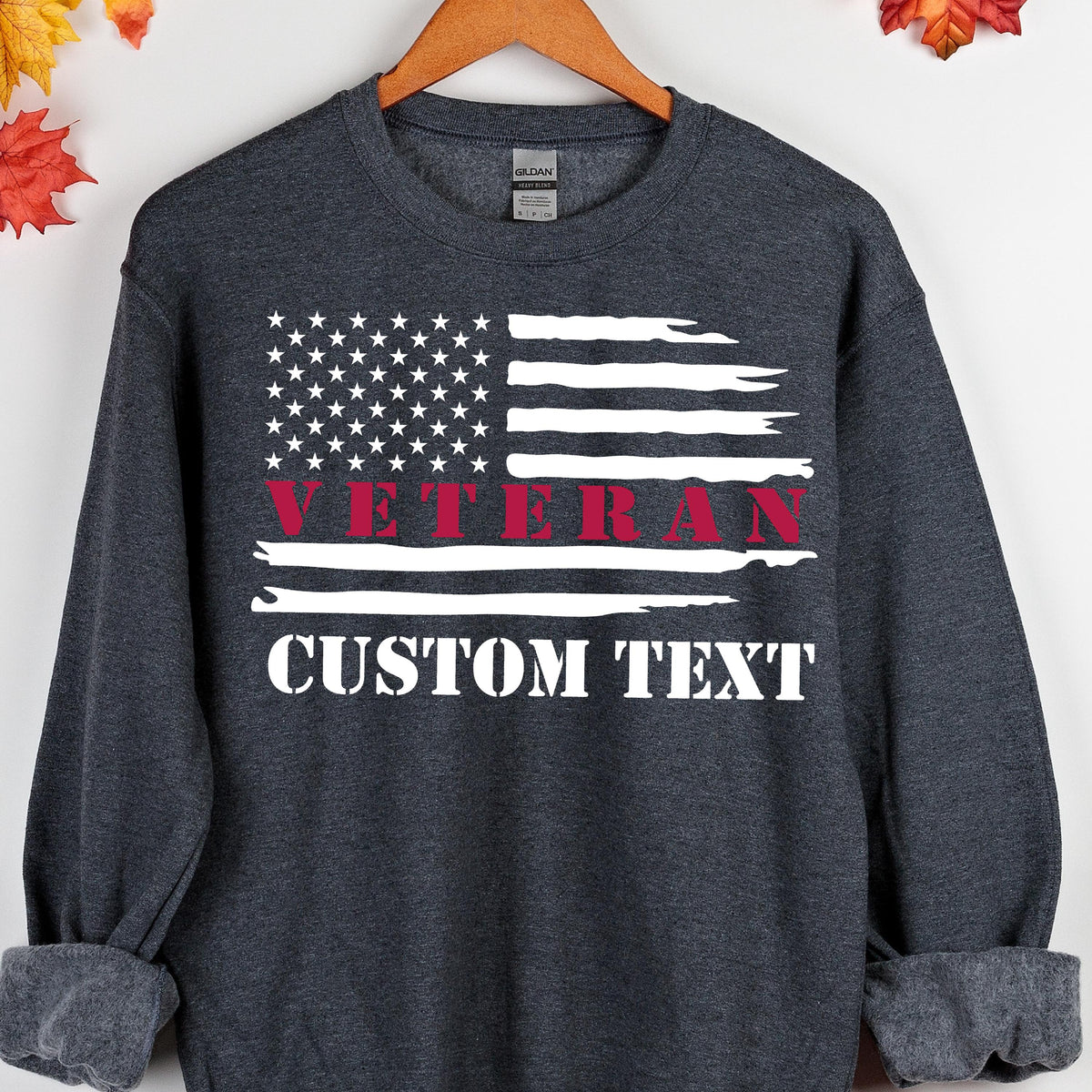 May include: Dark gray sweatshirt featuring a distressed American flag design. The flag includes white stars and stripes, with the word "VETERAN" in red and "CUSTOM TEXT" in white. The sweatshirt is displayed on a wooden hanger.