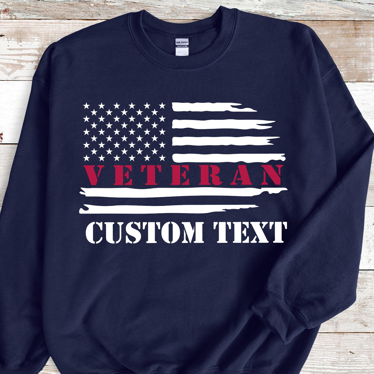 May include: Navy blue sweatshirt featuring a distressed American flag design. The flag includes white stars and stripes, with the word "VETERAN" in red and "CUSTOM TEXT" in white.