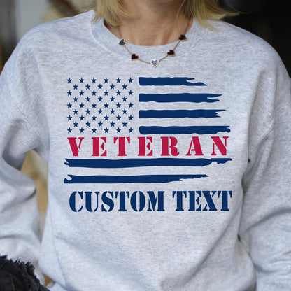 May include: A light gray sweatshirt featuring a distressed American flag design. The flag includes a field of stars, blue stripes, and the word "VETERAN" in red. Below the flag is the text "CUSTOM TEXT" in a bold, blue font.
