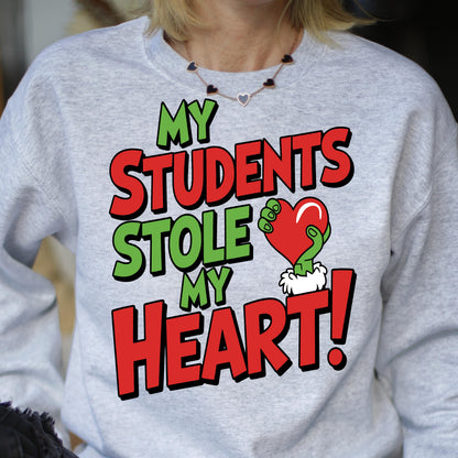 May include: A light gray sweatshirt with the text "MY STUDENTS STOLE MY HEART!" in green and red. The design includes a green hand holding a red heart. The sweatshirt is a crew neck style.