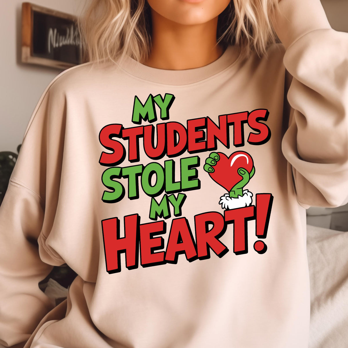 May include: Beige sweatshirt with the text "MY STUDENTS STOLE MY HEART!" in green and red. The design includes a green hand holding a red heart with white fur trim. A fun, teacher-themed graphic.