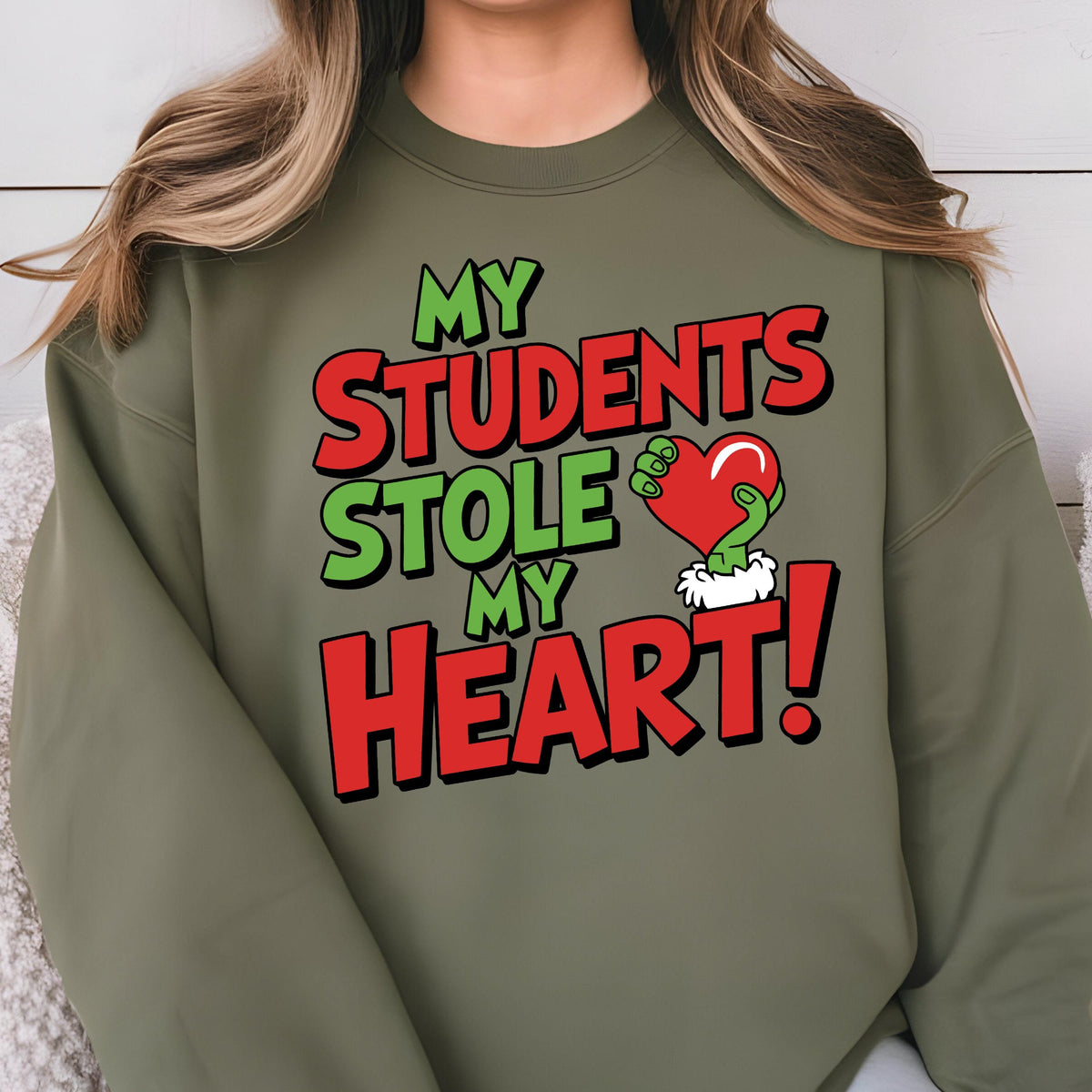 May include: Olive green sweatshirt with the text "MY STUDENTS STOLE MY HEART!" in red and green. The design includes a green hand holding a red heart. A fun and festive design for teachers.