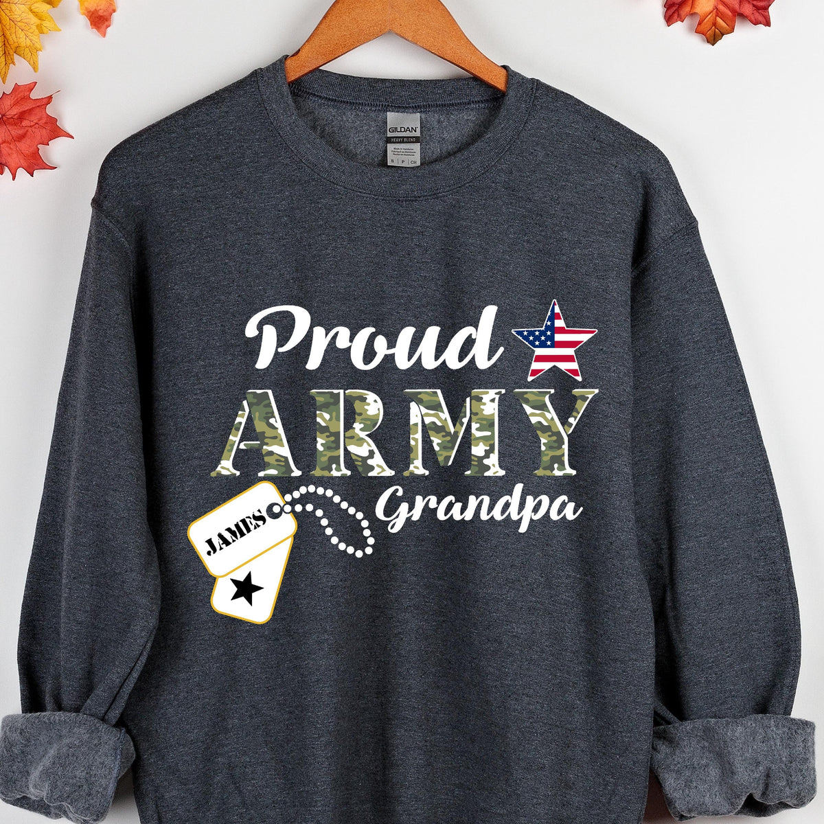 May include: Dark gray sweatshirt with the words "Proud ARMY Grandpa" in white script and camouflage lettering. A dog tag with the name "JAMES" is also visible. A red, white, and blue star graphic is on the right side of the sweatshirt.