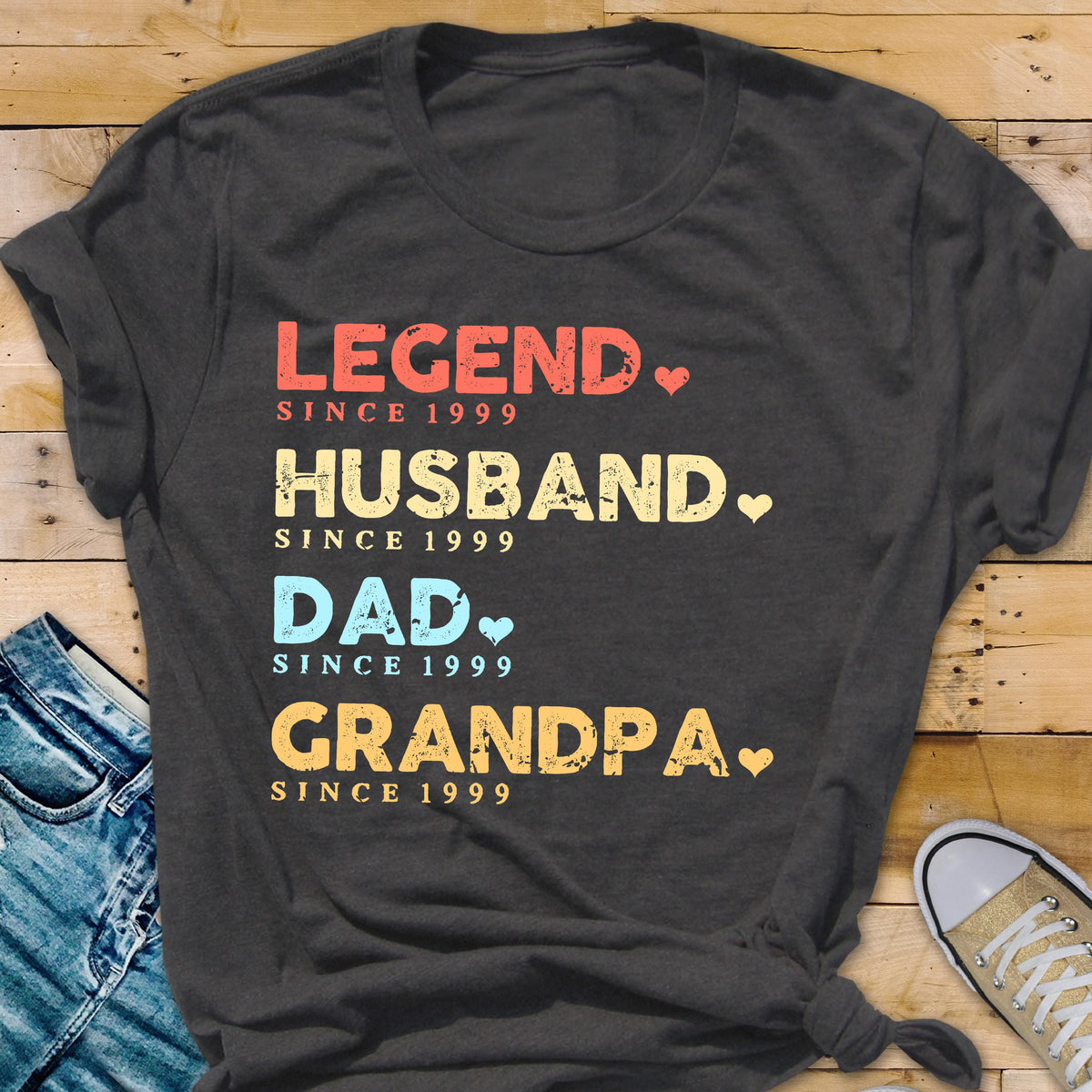May include: Dark gray t-shirt with the words "LEGEND, HUSBAND, DAD, GRANDPA" in red, yellow, blue, and orange text. Each word is followed by "SINCE 1999" and a heart symbol. A casual tee.
