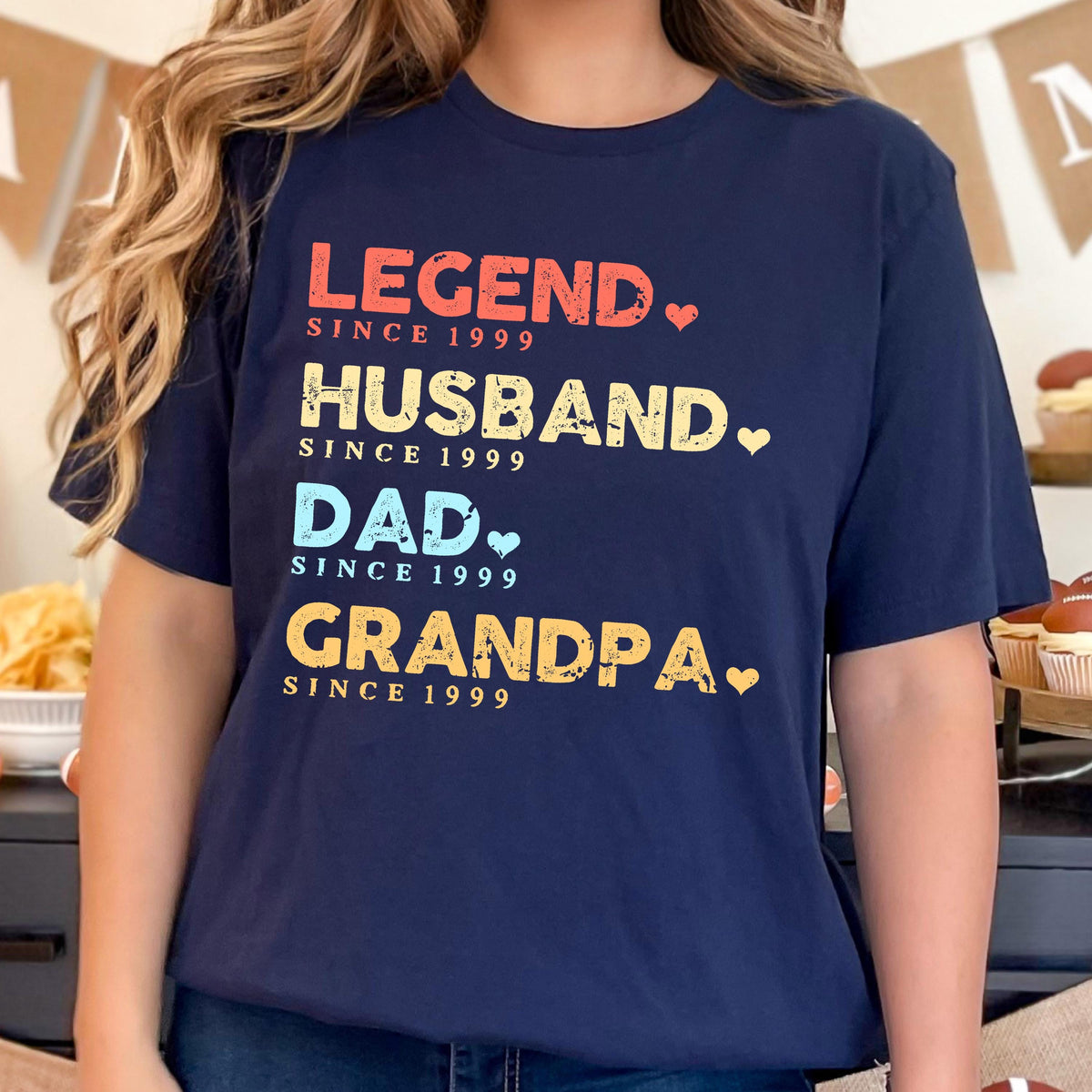 May include: Navy blue t-shirt with the words "LEGEND, HUSBAND, DAD, GRANDPA" in colorful text, each followed by "SINCE 1999". The design includes small heart shapes. A casual, family-themed apparel item.