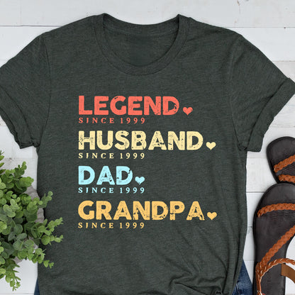 May include: Dark gray t-shirt with the words "LEGEND," "HUSBAND," "DAD," and "GRANDPA" in red, yellow, blue, and orange, respectively. Each word is followed by a heart and the text "SINCE 1999."