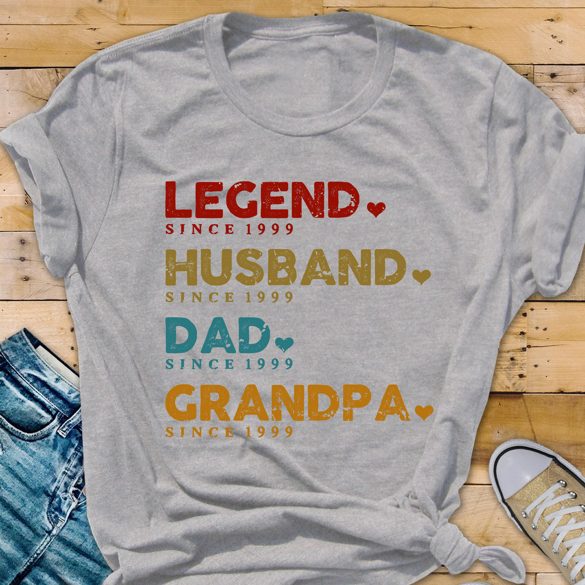 May include: A gray t-shirt with the words "LEGEND," "HUSBAND," "DAD," and "GRANDPA" printed in red, gold, blue, and orange, respectively. Each word is followed by "SINCE 1999." The shirt has a casual, everyday style.