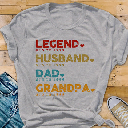 May include: A gray t-shirt with the words "LEGEND," "HUSBAND," "DAD," and "GRANDPA" printed in red, gold, blue, and orange, respectively. Each word is followed by "SINCE 1999." The shirt has a casual, everyday style.