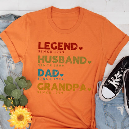May include: Orange t-shirt with the words "LEGEND, HUSBAND, DAD, GRANDPA" printed in various colors. Each word is followed by "SINCE 1999". The shirt is folded, with a denim jacket, sneakers, and a flower visible in the background.