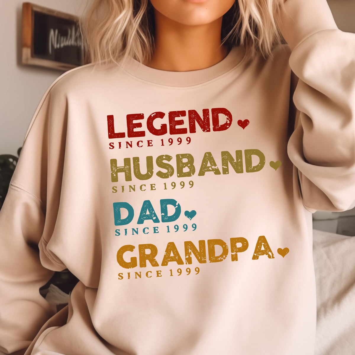 May include: A beige sweatshirt with the words "LEGEND," "HUSBAND," "DAD," and "GRANDPA" in various colors. Each word is followed by "SINCE 1999." The design includes small heart accents, creating a personalized gift.