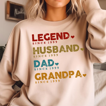 May include: A beige sweatshirt with the words "LEGEND," "HUSBAND," "DAD," and "GRANDPA" in various colors. Each word is followed by "SINCE 1999." The design includes small heart accents, creating a personalized gift.