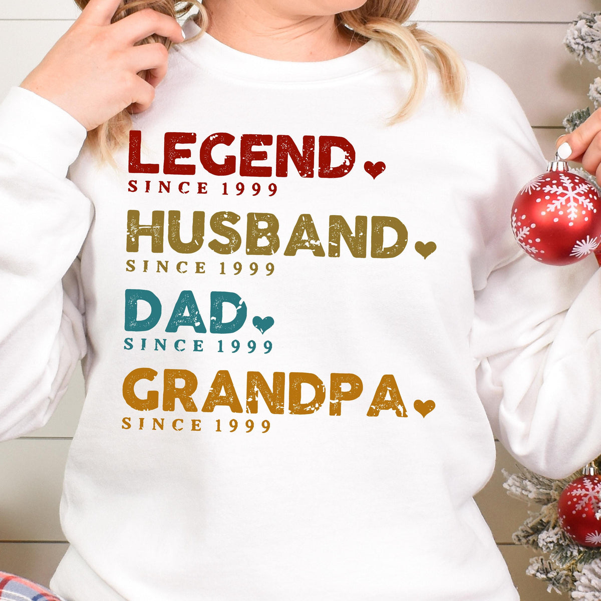 May include: White sweatshirt with the words "LEGEND," "HUSBAND," "DAD," and "GRANDPA" in various colors. Each word is followed by "SINCE 1999." A heart graphic is next to each word. A festive holiday ornament is in the background.