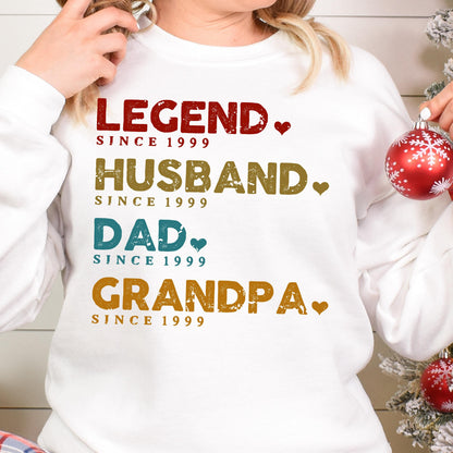 May include: White sweatshirt with the words "LEGEND," "HUSBAND," "DAD," and "GRANDPA" in various colors. Each word is followed by "SINCE 1999." A heart graphic is next to each word. A festive holiday ornament is in the background.