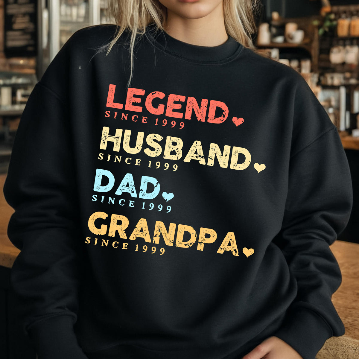 May include: Black sweatshirt with the words "LEGEND, HUSBAND, DAD, GRANDPA" in red, yellow, blue, and orange text. Each word is followed by "SINCE 1999" and a heart symbol. The sweatshirt is a crew neck style.