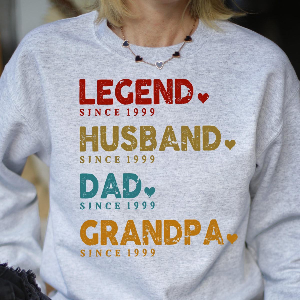 May include: A light gray sweatshirt with the words "LEGEND," "HUSBAND," "DAD," and "GRANDPA" printed in red, gold, teal, and orange, respectively. Each word is followed by a heart and the phrase "SINCE 1999."