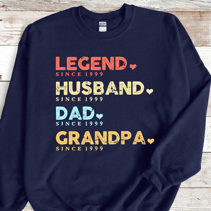 May include: Navy blue sweatshirt with the words "LEGEND, HUSBAND, DAD, GRANDPA" in various colors, each followed by "SINCE 1999". The text has a heart symbol next to each word. A comfortable, casual top.
