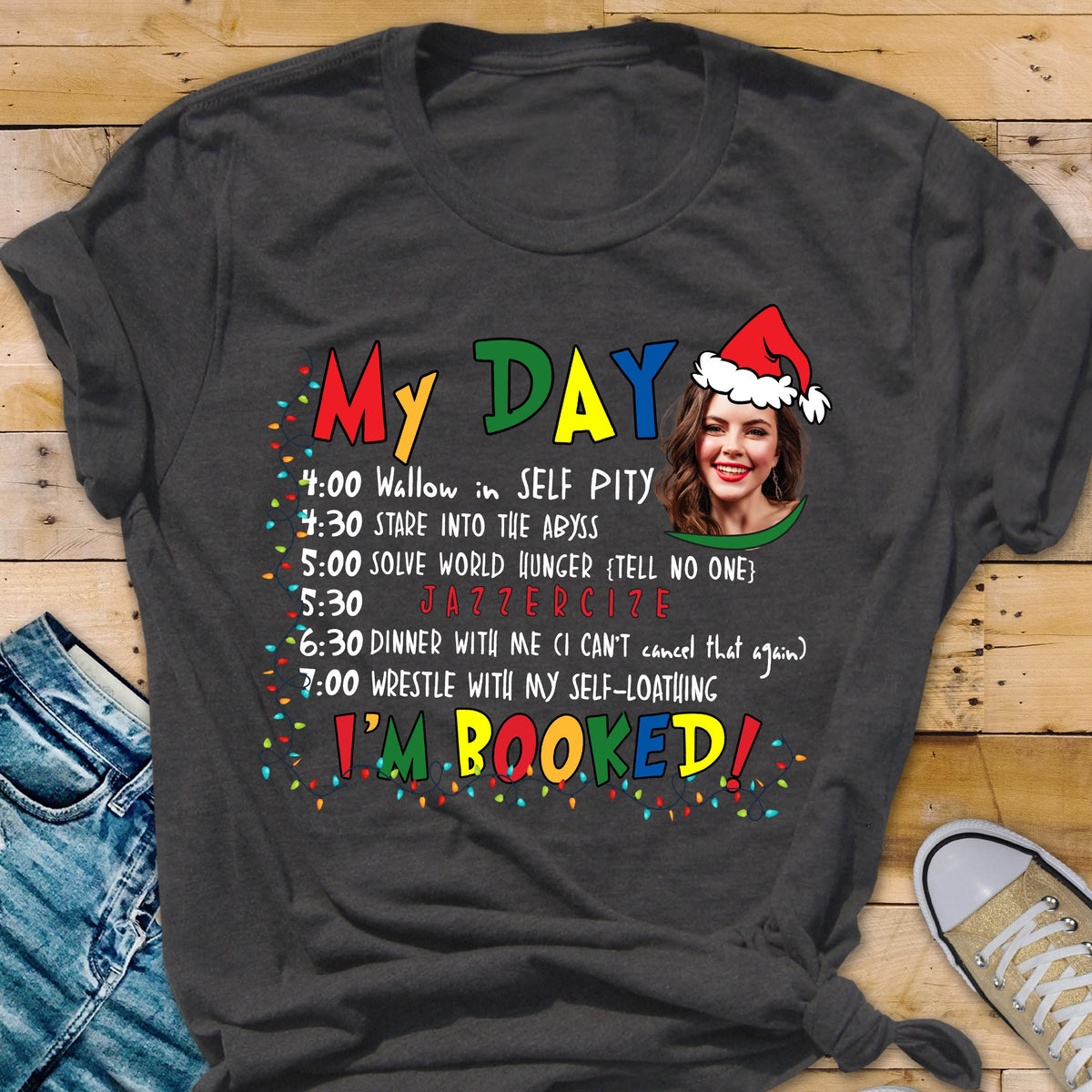 May include: Dark gray t-shirt with the words "My DAY" in colorful letters. The shirt features a schedule of humorous activities, including "Wallow in SELF PITY" and "JAZZERCIZE." A woman wearing a Santa hat is pictured. The text "I'M BOOKED!" is also displayed.