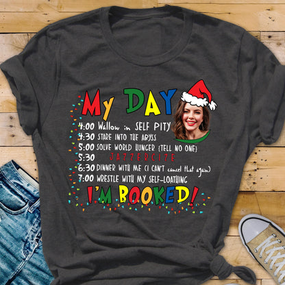 May include: Dark gray t-shirt with the words "My DAY" in colorful letters. The shirt features a schedule of humorous activities, including "Wallow in SELF PITY" and "JAZZERCIZE." A woman wearing a Santa hat is pictured. The text "I'M BOOKED!" is also displayed.