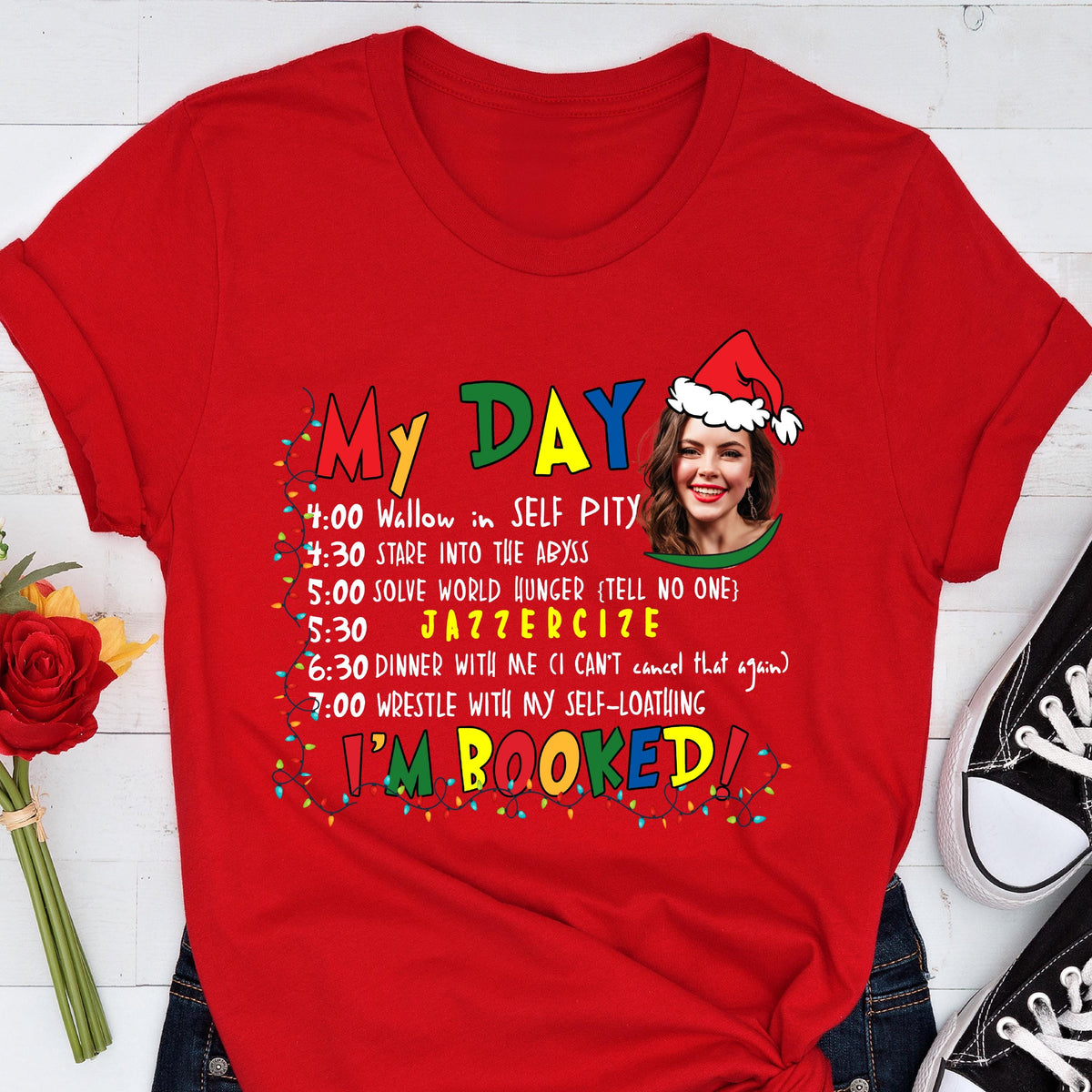 May include: Red t-shirt with the text "My Day" and a humorous schedule including "Wallow in Self Pity" and "Jazzercise." A woman wearing a Santa hat is in the design. The words "I'm Booked!" are also on the shirt.
