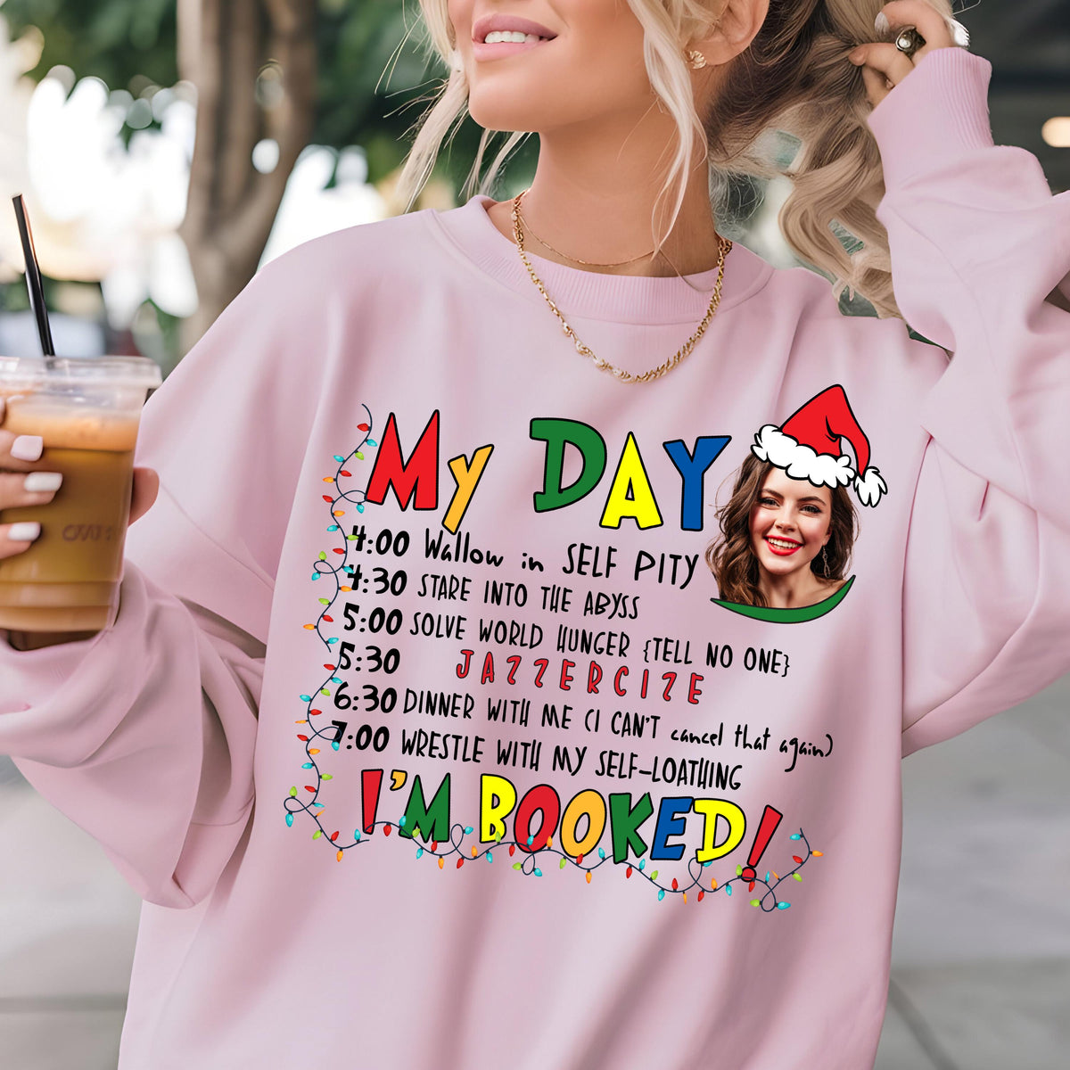 May include: A light pink crewneck sweatshirt featuring a graphic print with the text My Day followed by a schedule of activities, a photo of a person wearing a Santa hat, and the text I'm Booked, all framed by colorful Christmas light strings.