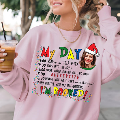 May include: A light pink crewneck sweatshirt featuring a graphic print with the text My Day followed by a schedule of activities, a photo of a person wearing a Santa hat, and the text I'm Booked, all framed by colorful Christmas light strings.