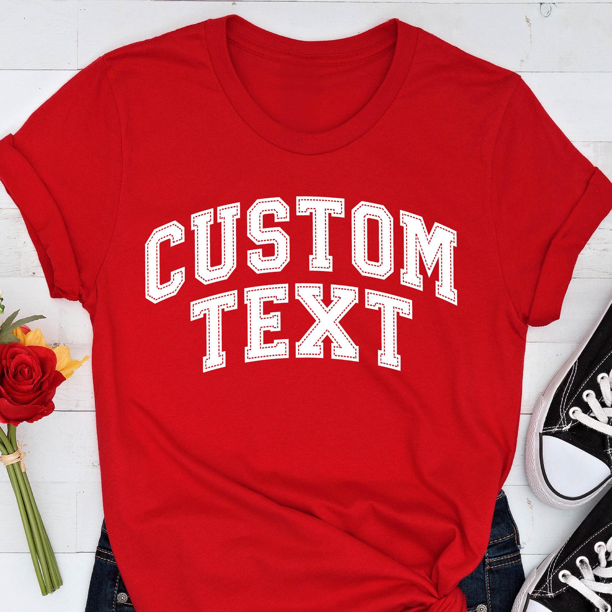 May include: A red t-shirt with the words "CUSTOM TEXT" in white, collegiate-style lettering. The text has a stitched outline. The shirt is paired with blue jeans and black sneakers.