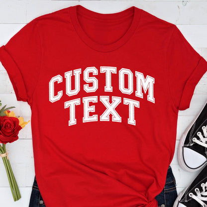 May include: A red t-shirt with the words "CUSTOM TEXT" in white, collegiate-style lettering. The text has a stitched outline. The shirt is paired with blue jeans and black sneakers.