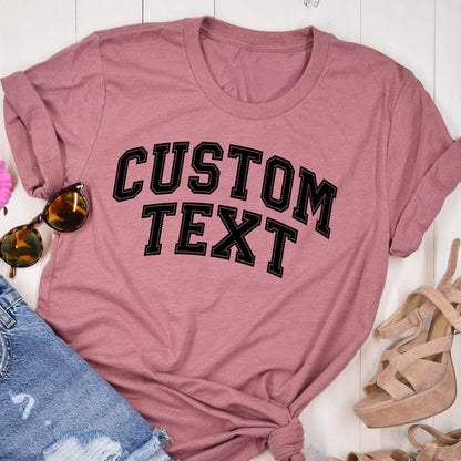 May include: A dusty rose-colored t-shirt with the words "CUSTOM TEXT" in a black, collegiate-style font. The text is outlined in white stitching. The shirt is styled with sunglasses, denim shorts, and platform sandals.