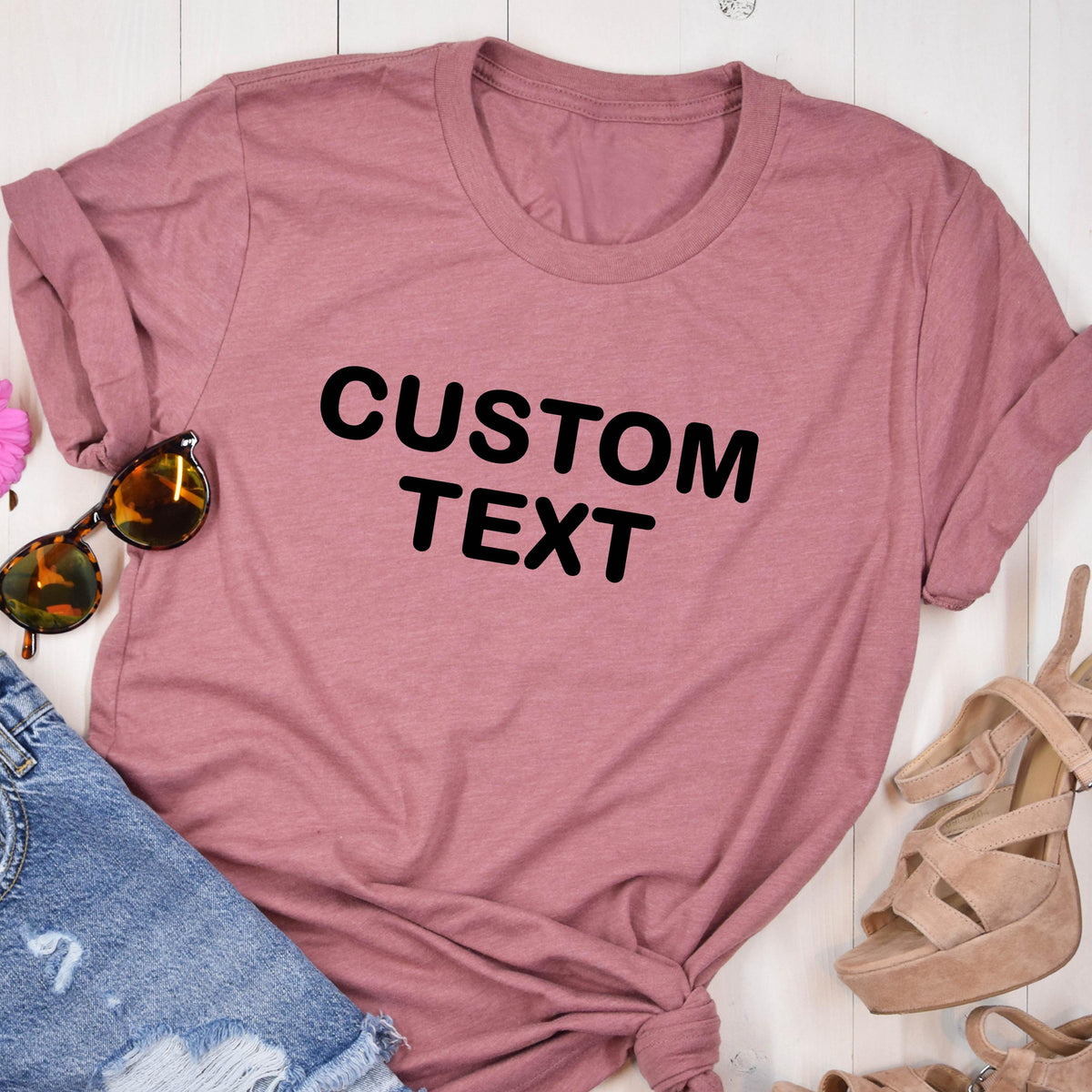 May include: A dusty rose short-sleeved crew neck t-shirt featuring the words CUSTOM TEXT printed in bold black capital letters across the center of the chest, paired with blue denim fabric and tan high-heeled sandals with sunglasses.