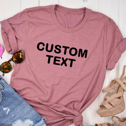 May include: A dusty rose short-sleeved crew neck t-shirt featuring the words CUSTOM TEXT printed in bold black capital letters across the center of the chest, paired with blue denim fabric and tan high-heeled sandals with sunglasses.