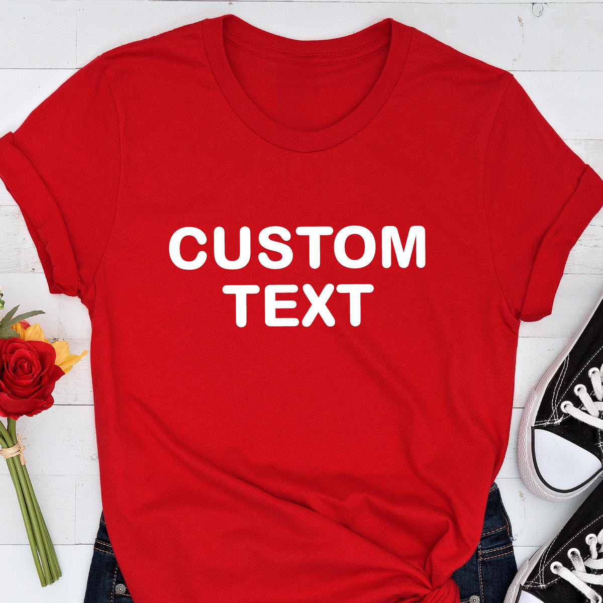 May include: A bright red short-sleeved t-shirt featuring the words CUSTOM TEXT printed in bold white capital letters across the center of the chest. The shirt has a classic crew neckline and is displayed flat with rolled sleeves.