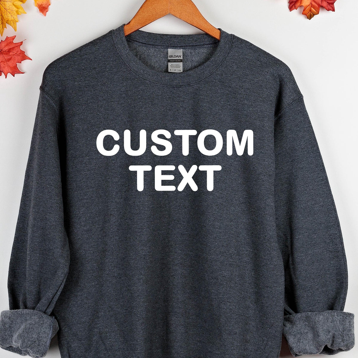 May include: A dark heather gray crewneck sweatshirt featuring the white text CUSTOM TEXT printed in a bold sans-serif font across the chest. The garment has long sleeves with ribbed cuffs and a ribbed neckline, displayed on a wooden hanger.