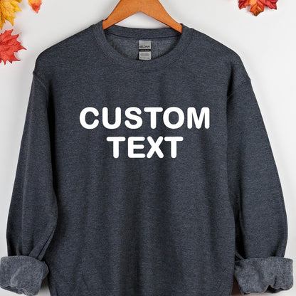 May include: A dark heather gray crewneck sweatshirt featuring the white text CUSTOM TEXT printed in a bold sans-serif font across the chest. The garment has long sleeves with ribbed cuffs and a ribbed neckline, displayed on a wooden hanger.