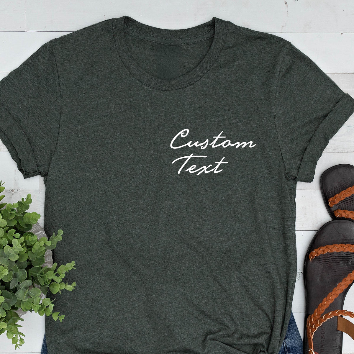 May include: Dark gray t-shirt with rolled sleeves. The shirt features the white cursive text "Custom Text" on the upper left chest. The shirt is paired with blue jeans and brown sandals.