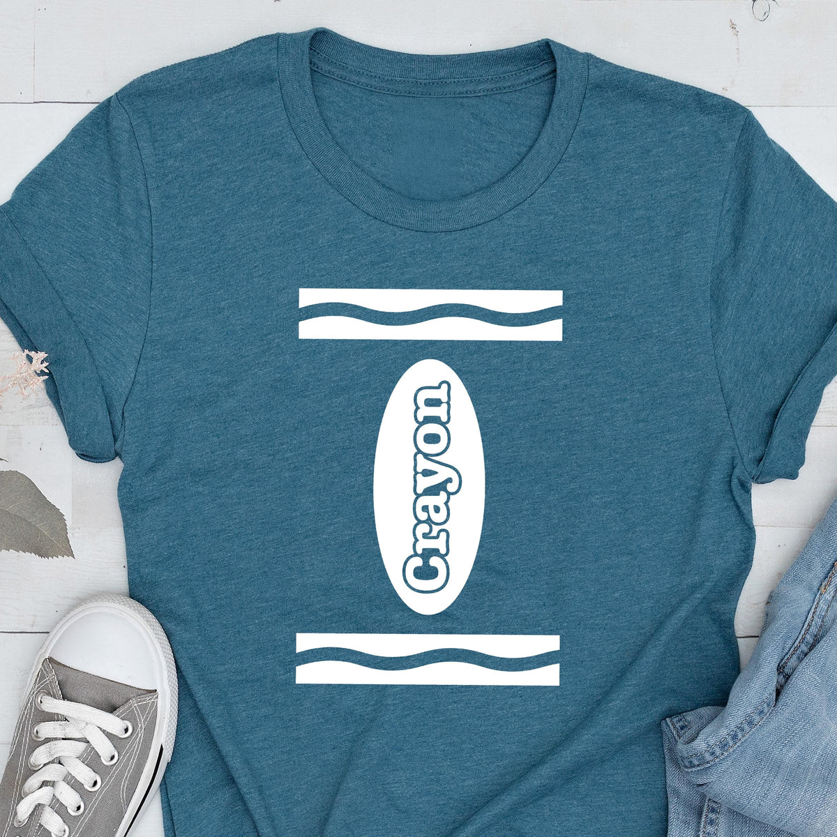 May include: A teal blue t-shirt with a white crayon design. The design features a white oval with the word "Crayon" written inside. The oval is surrounded by two white wavy lines.