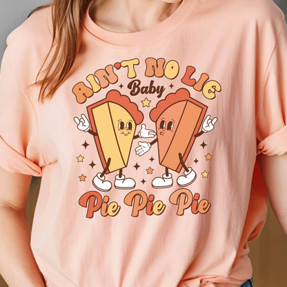 May include: Peach-colored t-shirt featuring a retro graphic of two animated pie slices with arms and legs. The text "Ain't No Lie Baby" and "Pie Pie Pie" are in a vintage font. The design includes stars.
