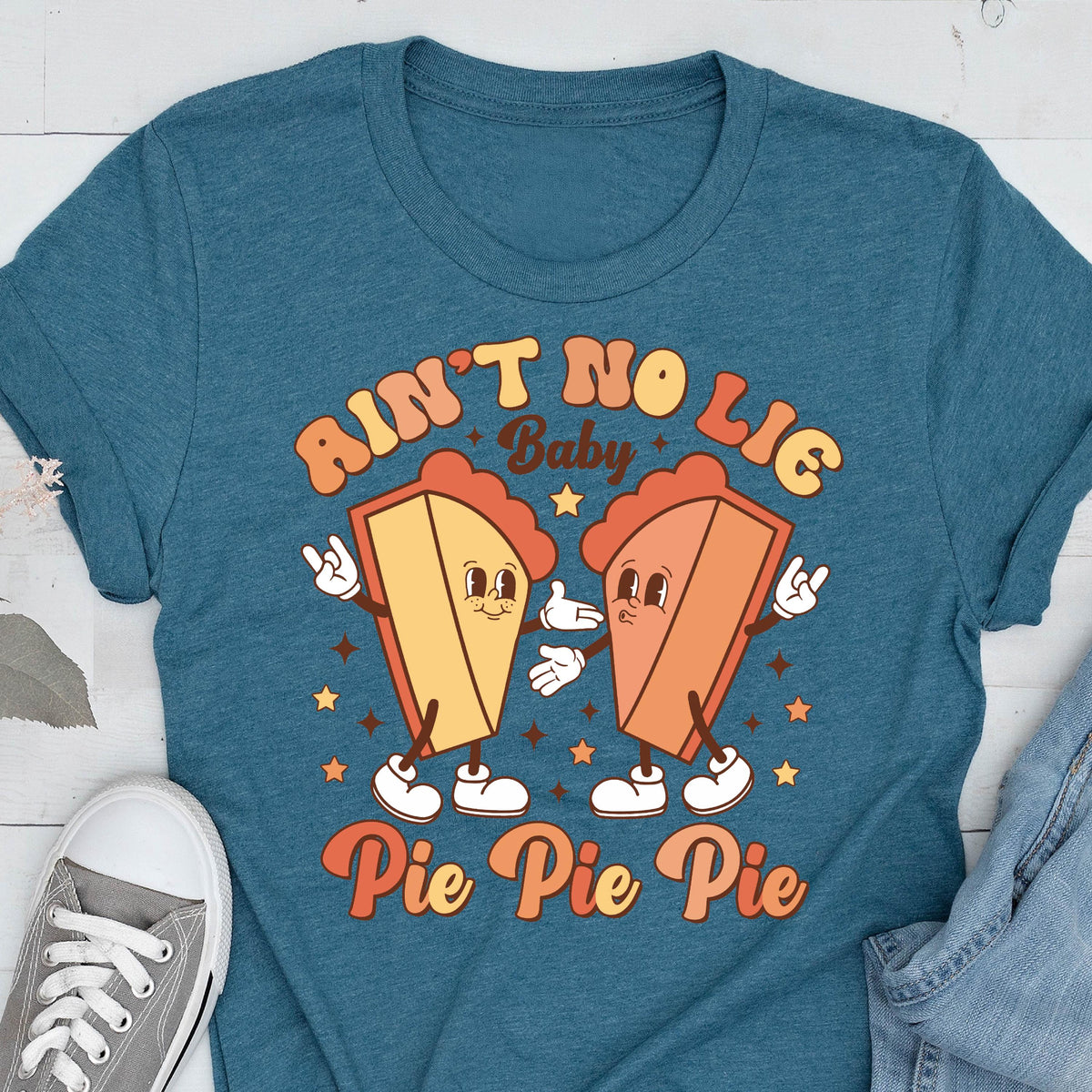 May include: Teal t-shirt featuring a retro graphic with two cartoon pie slices with arms and legs. The text reads "Ain't No Lie Baby" above the pies and "Pie Pie Pie" below. The design has a vintage, groovy aesthetic.