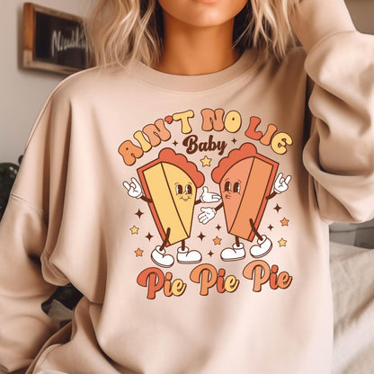 May include: A beige crewneck sweatshirt featuring a graphic print of two anthropomorphic pie slices with arms and legs. The text reads AIN'T NO LIE Baby Pie Pie Pie in orange and yellow tones, surrounded by small star shapes on the front chest.