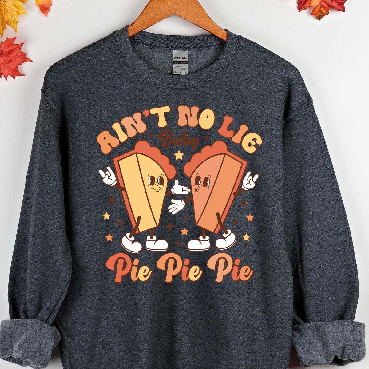May include: A dark charcoal gray crewneck sweatshirt featuring a graphic print of two anthropomorphic pie slices with arms and legs. The text reads "AIN'T NO LIE" and "Pie Pie Pie" in a retro font, surrounded by small orange stars on the front.
